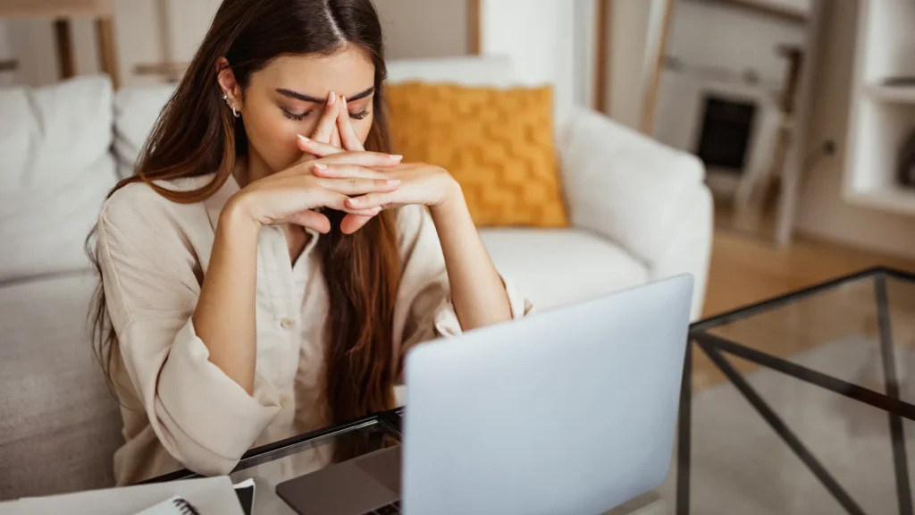 La chica frente al portátil se sostiene la frente con los ojos cerrados, en estado de cansancio.