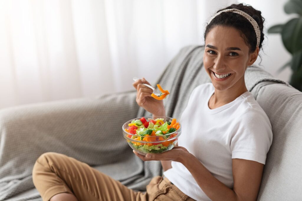 Donna che mangia insalata sul divano di casa.