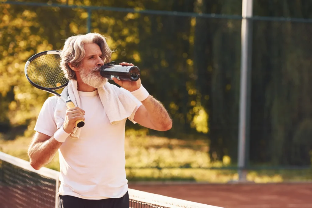 Elegante uomo di mezza età beve acqua dalla sua borraccia durante la pausa di una partita di tennis