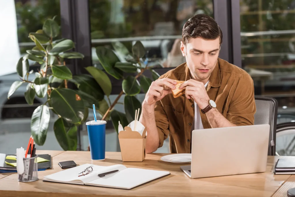 Ragazzo che mangia un tramezzino alla sua scrivania, mentre lavora al pc