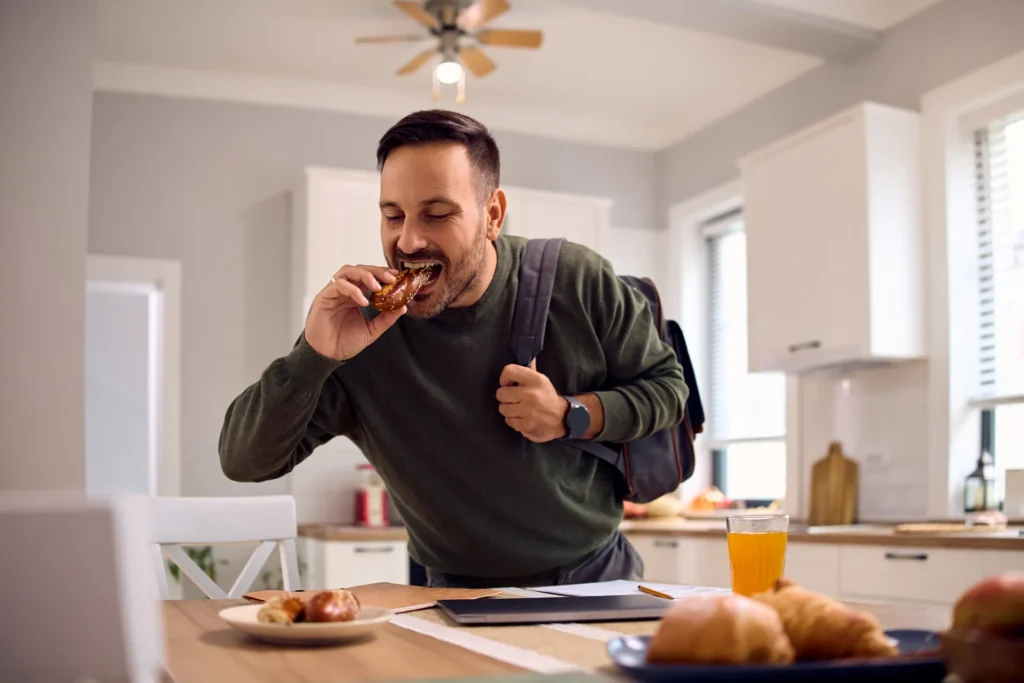 Uomo in cucina che mangia rapidamente un dolce mentre indossa uno zaino, con un tavolo apparecchiato per la colazione davanti a lui.