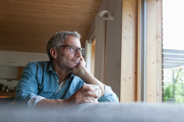 Un uomo di mezza età seduto vicino alla finestra, con una tazza in mano e lo sguardo pensieroso rivolto verso l’esterno.