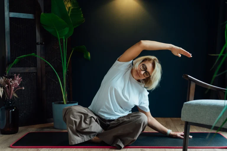 Donna di mezza età seduta su un tappetino da yoga in casa mentre esegue un esercizio di stretching laterale; accanto a lei una pianta in vaso e una sedia.