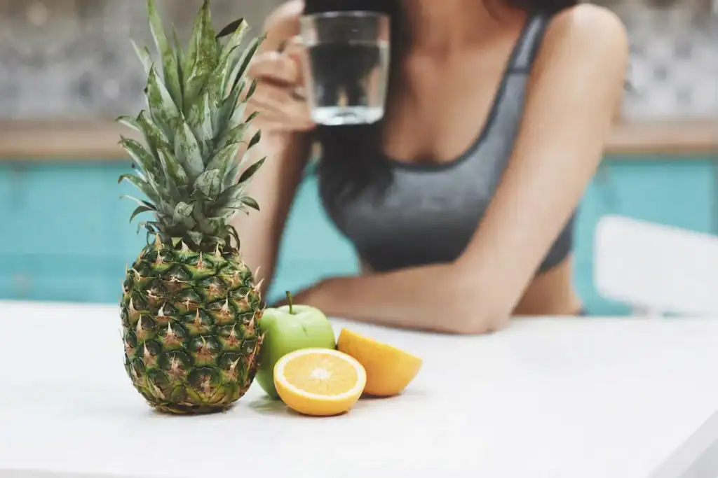 In primo piano: un ananas intero, una mela verde e due mezzi limoni appoggiati su un tavolo bianco. Sullo sfondo, sfocato, una persona con un top grigio tiene in mano un bicchiere d'acqua.
