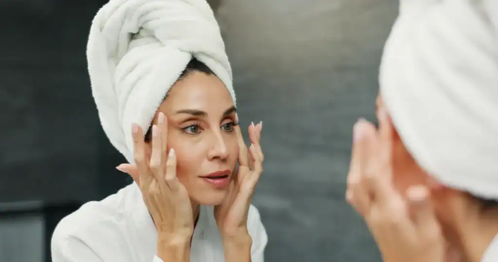 Donna con un asciugamano bianco sulla testa che si osserva il viso allo specchio toccando delicatamente la pelle con entrambe le mani.