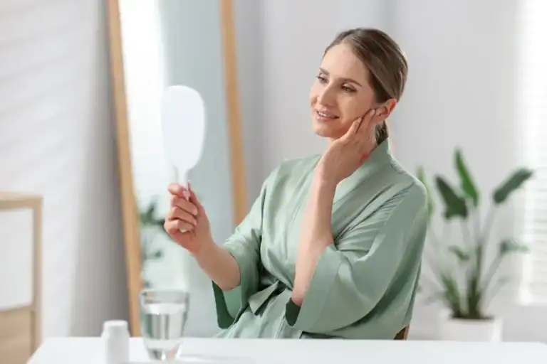 Donna con un accappatoio verde che tiene uno specchio in mano e si tocca la guancia mentre osserva il proprio viso, seduta davanti a un tavolo con un bicchiere d'acqua.