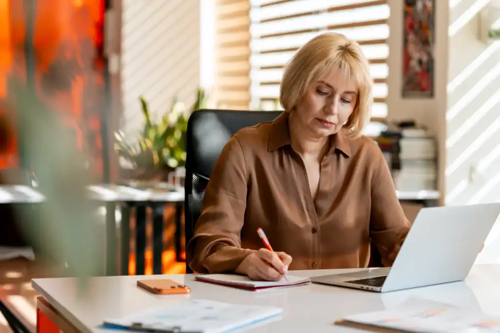 Donna adulta seduta alla scrivania mentre prende appunti su un quaderno e lavora al computer portatile.