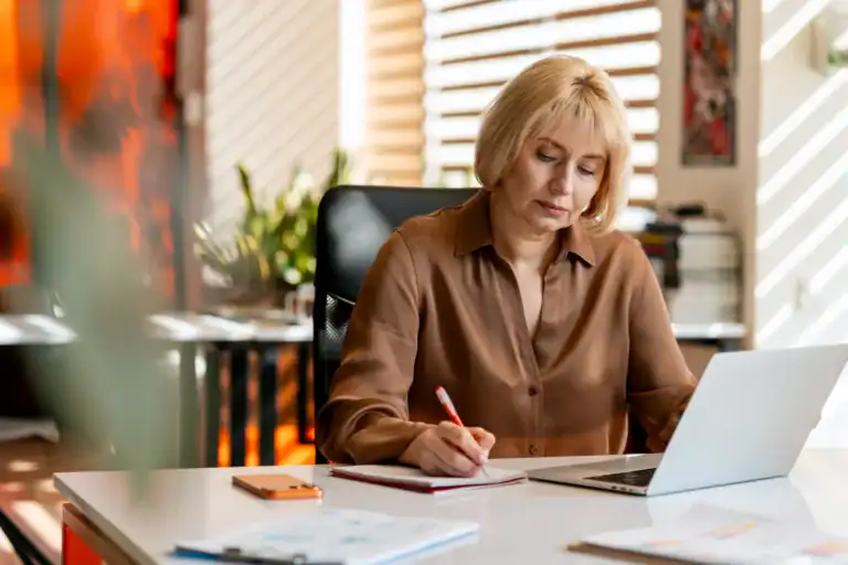 Donna adulta seduta alla scrivania mentre prende appunti su un quaderno e lavora al computer portatile.